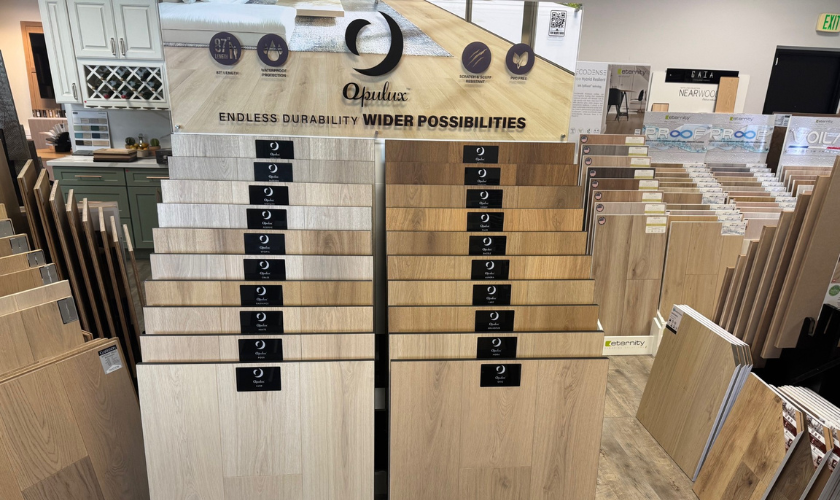 Opulux waterproof flooring display in a showroom with tiered wood-look planks labeled by style. Features include waterproof, scratch-resistant, pet-friendly, and PVC-free. Certifications like FloorScore and GreenGuard are shown. Background includes cabinetry and competing brand displays.